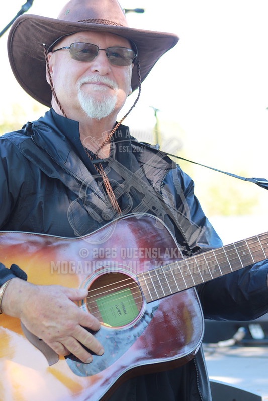 Jeff Creech smiling warmly in cowboy hat and glasses with sunburst acoustic guitar on stage at Queenslake