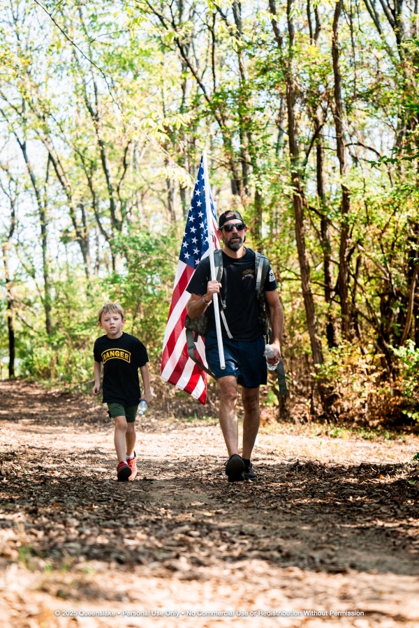 Veteran and Son Walking Along Sunlit Wooded Trail with Flag and Ruck