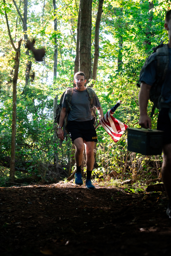 2026 Cadet with American Flag and Combat Ruck Powering Through Wooded Canopy